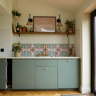 Green/blue cupboards, gold leaf island unit, large patterned tiles above the sink. Wooden dining table, with wooden slatted wall behind. Utility behind dining table and snug area behind kitchen