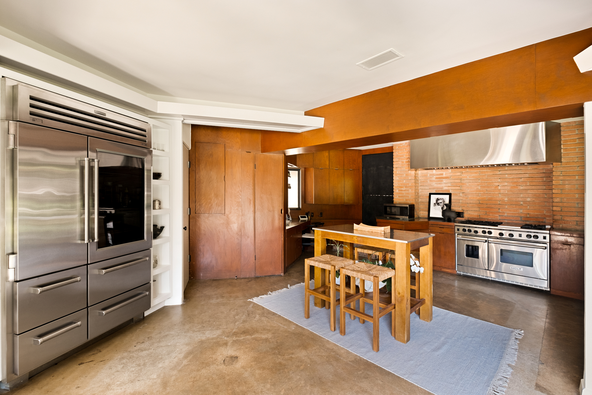 Image of a kitchen with white walls, warm wooden cabinetry and ceiling beams, concrete floors, brick backsplash, and stainless steel appliances.