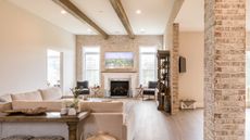 Bright, open living room area with brick walls and pillars, wooden floor, wooden ceiling beams and windows revealing bright outdoors. Pictured inside are a cream sofa, brown side table, plants and misc ornaments.