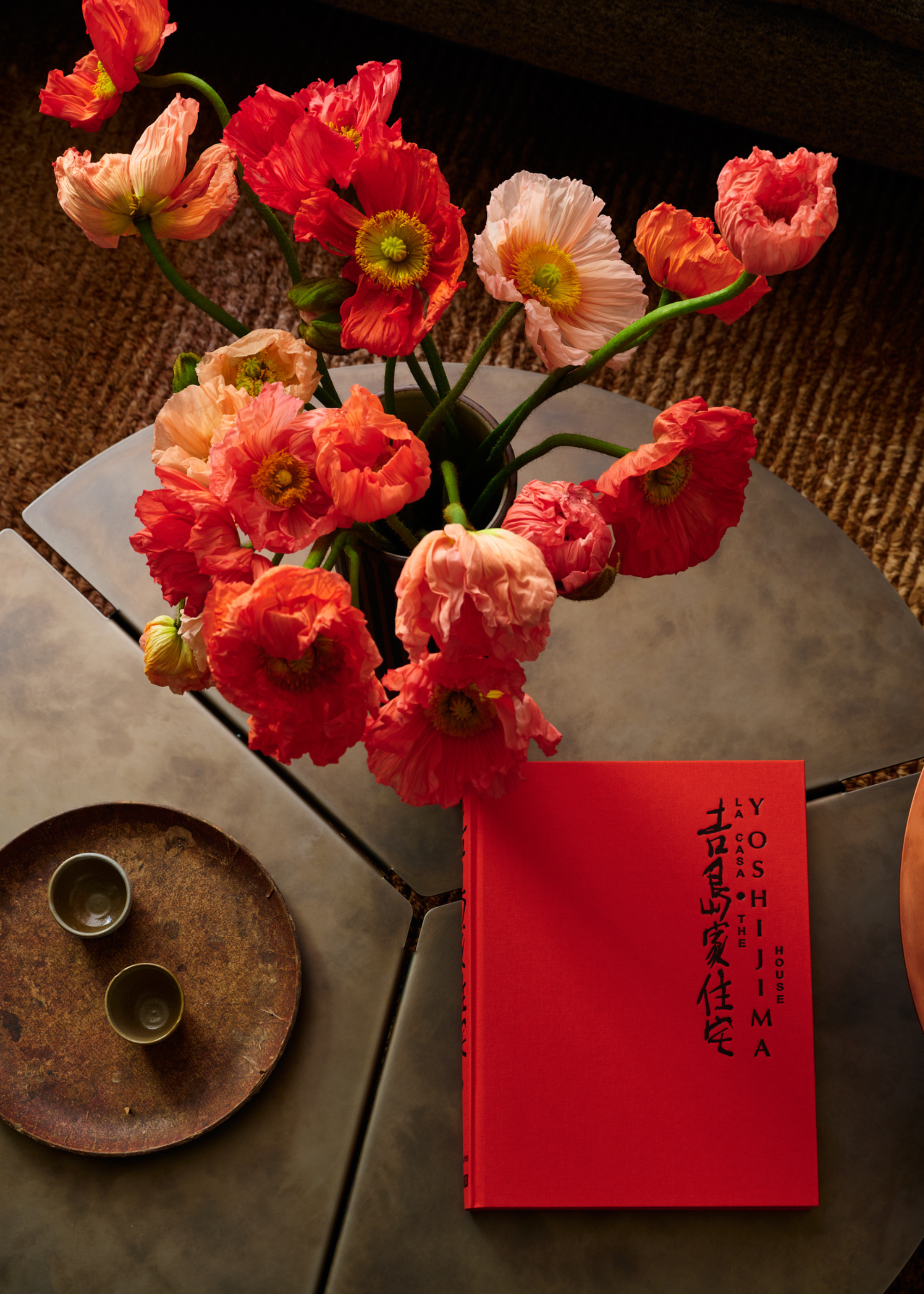 An overhead view of a coffee table with a vase of colorful flowers by a red book and a ceramic bowl