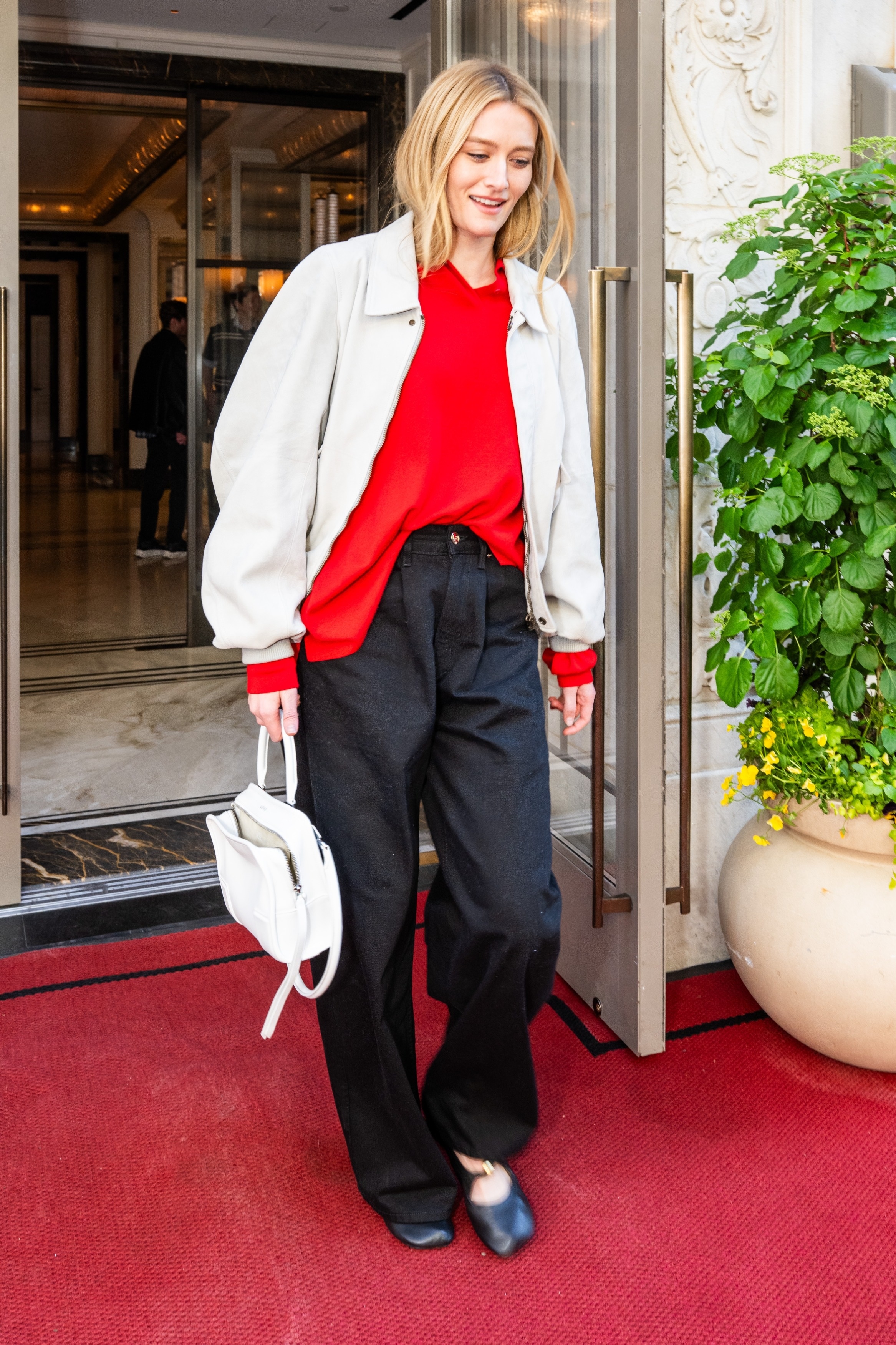 Sarah Pidgeon steps outside of a hotel wearing a red jumper with a beige jacket and black trousers.