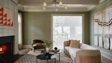 living room with olive green grasscloth wallpaper, oranate trim in white, pendnat light, large artwork on back wall, fireplace with wall molding, beige sofa with cane backing, two black round nesting tables, and a brown armchair