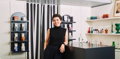 A young woman dressed in a sleeveless black turtleneck smiles at the camera in a design showroom filled with candles and glassware creations.