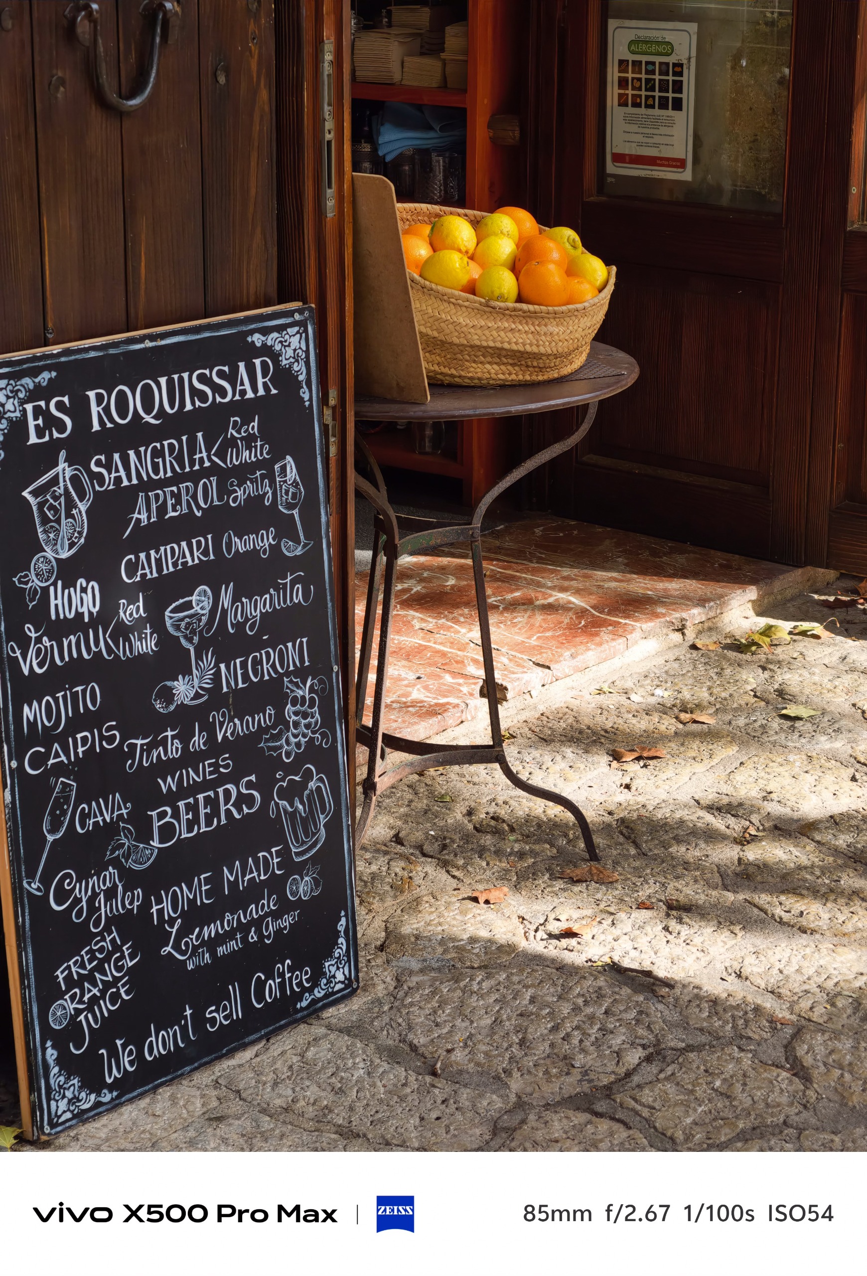 Chalkboard drinks menu beside a woven basket filled with oranges and lemons outside a rustic bar.