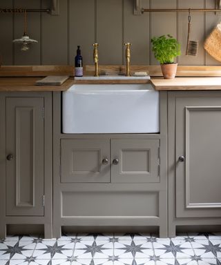 A farmhouse-style kitchen sink area with a white ceramic apron-front sink set against sage green cabinetry and oak countertops. The backdrop features vertical tongue-and-groove panelling, brass taps, and a pot of fresh herbs on the counter.