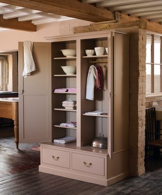 Pale pink painted cabinet with open doors, casserole dishes, linens, original wooden floorboards, beams