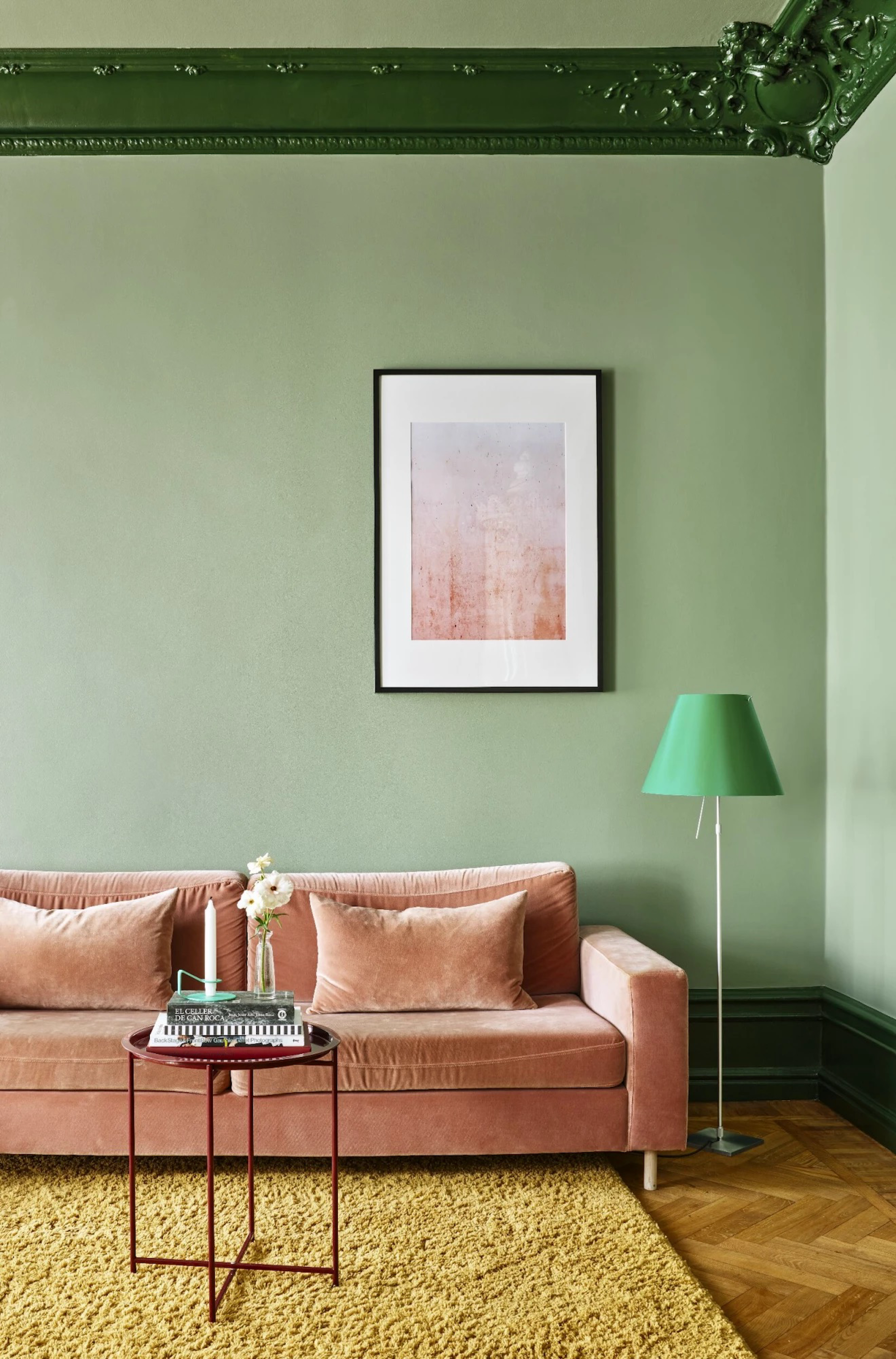 An avocado green living space with forrest green cornicing, a peach sofa, mustard rug underneath and an artwork on the wall behind the sofa.