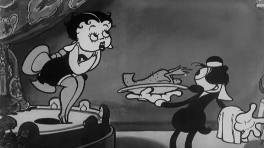 A black-and-white still from an animated cartoon shows Betty Boop standing on a bed, looking down at Pudgy (a dog character) who is dressed as a waiter and presenting a platter with a roasted bird.