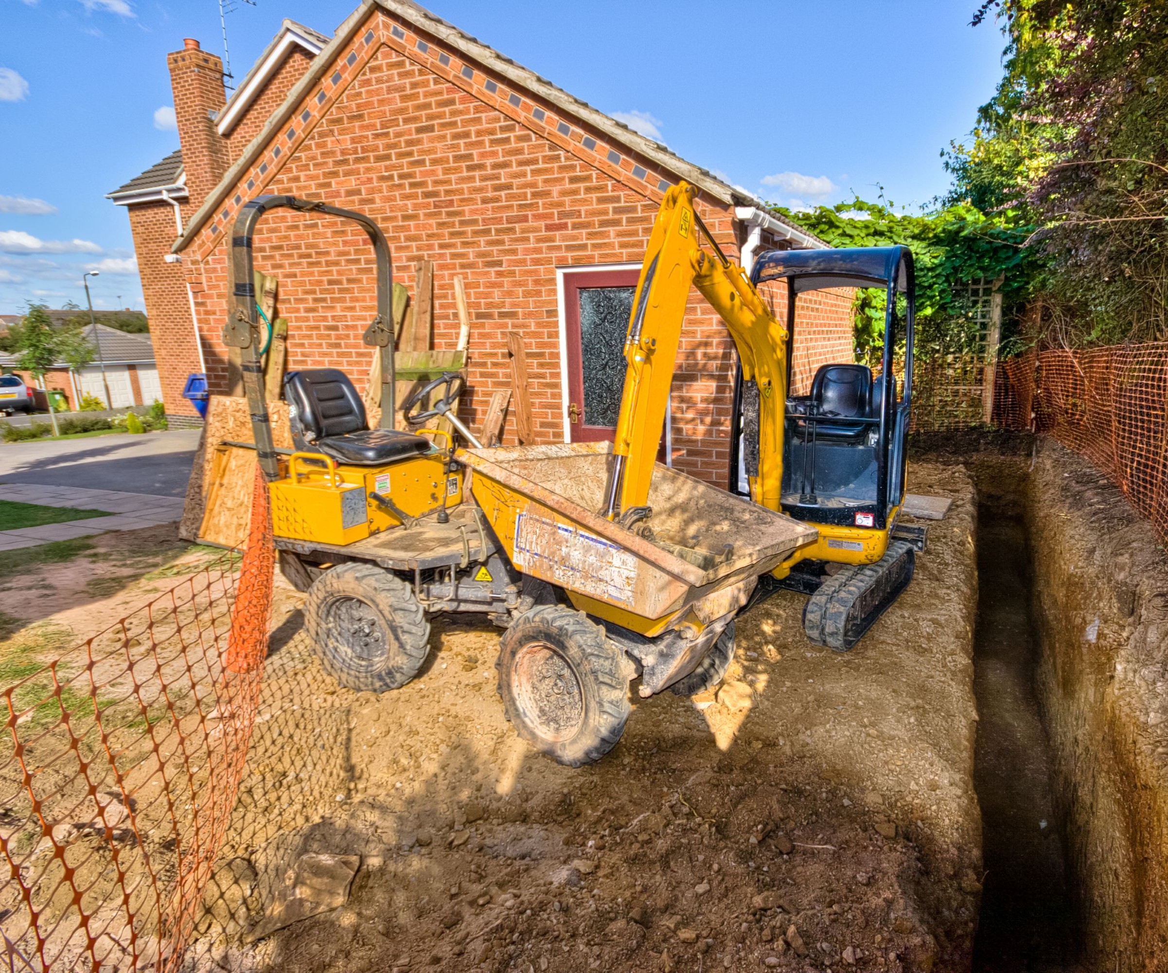 A yellow digger and yellow dumper excavating a large trench in front of a red brick bungalow