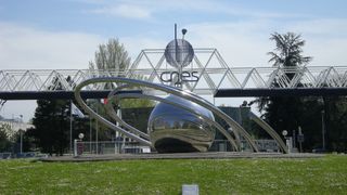 A metal ball surrounded by metal rings sits on a grassy lawn in front of a glass building with the letters CNES on the front