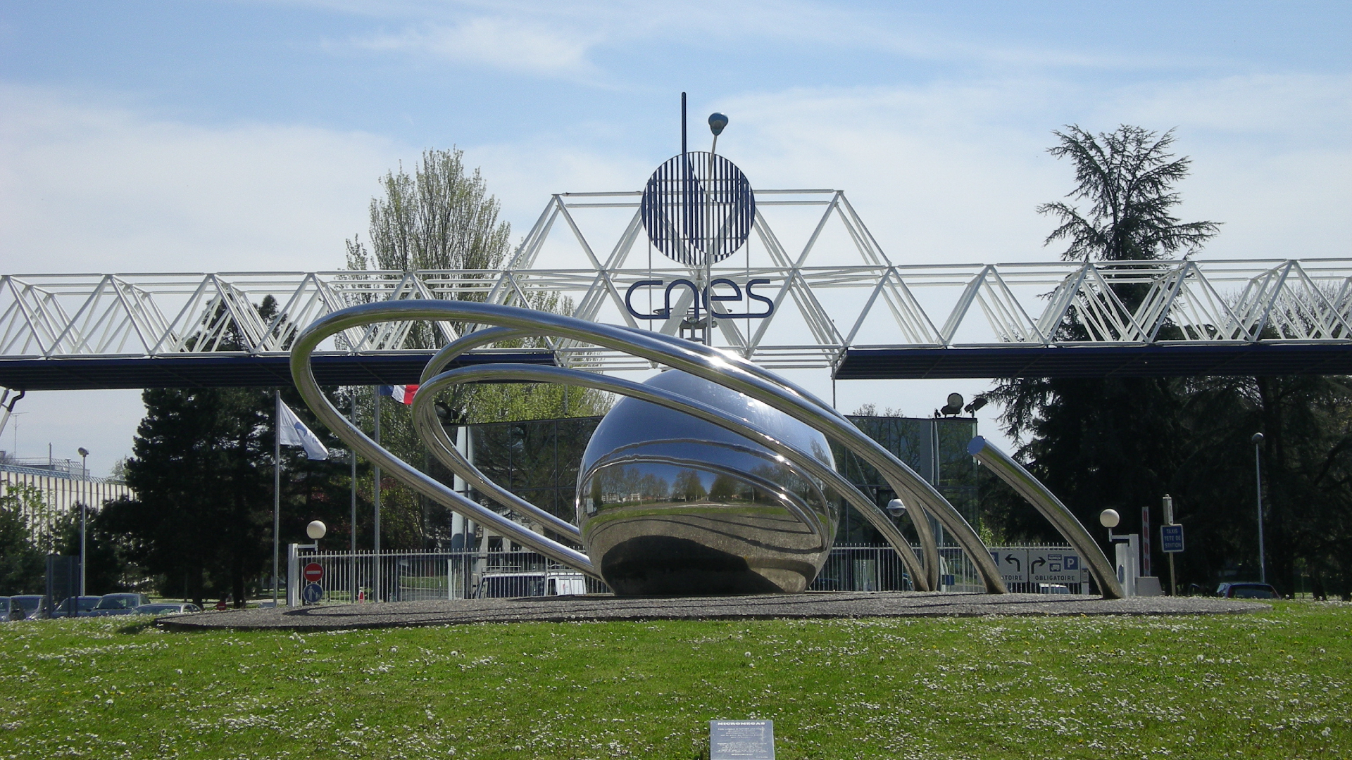 A metal ball surrounded by metal rings sits on a grassy lawn in front of a glass building with the letters CNES on the front