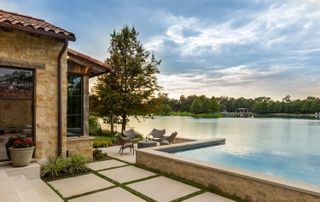 lakeside exterior of stone built house with trees and patio loungers