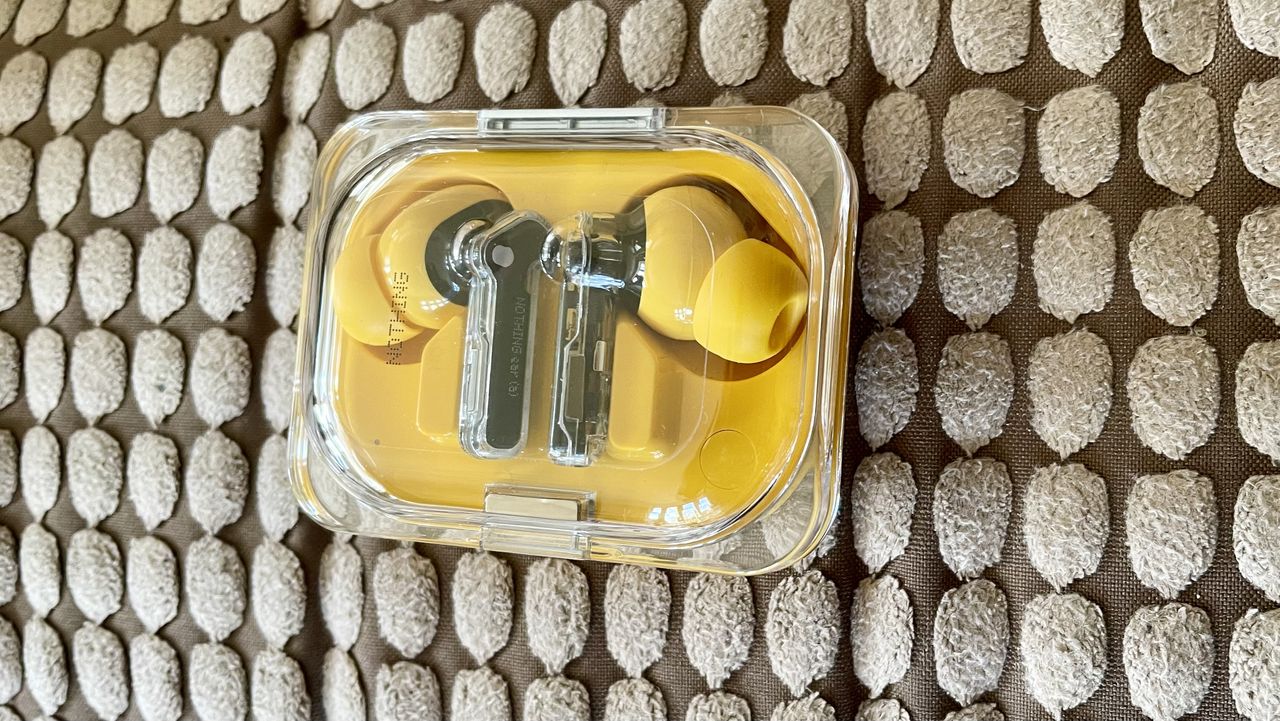 Nothing Ear (a) earbuds on a brown sofa, in their closed case &ndash;&nbsp;with transparent lid