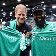 Prince Harry and a man smiling and holding green jerseys that say Lilibet and Archie in a basketball arena