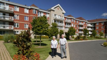 An older couple walk outside a senior housing facility on a sunny day.
