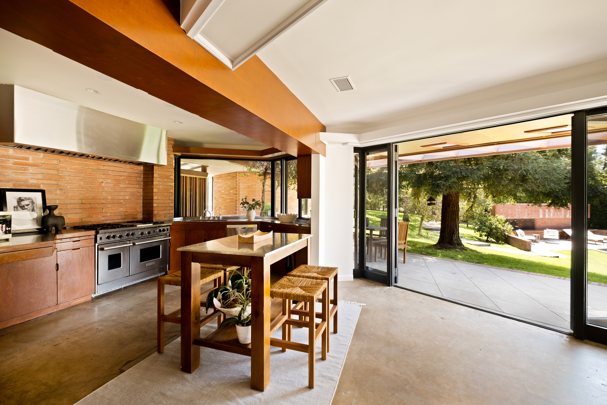 Image of a kitchen with white walls, warm wooden cabinetry and ceiling beams, concrete floors, brick backsplash, and stainless steel appliances. There is a small island dining table in the center and a windowed door that is open to outside.