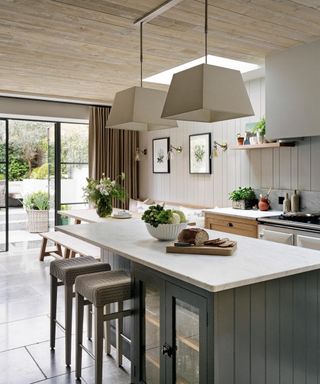 Neutral kitchen with wooden panelled ceiling, paneling on walls, large square pendant lights, kitchen island, dining area, artwork, wall lights