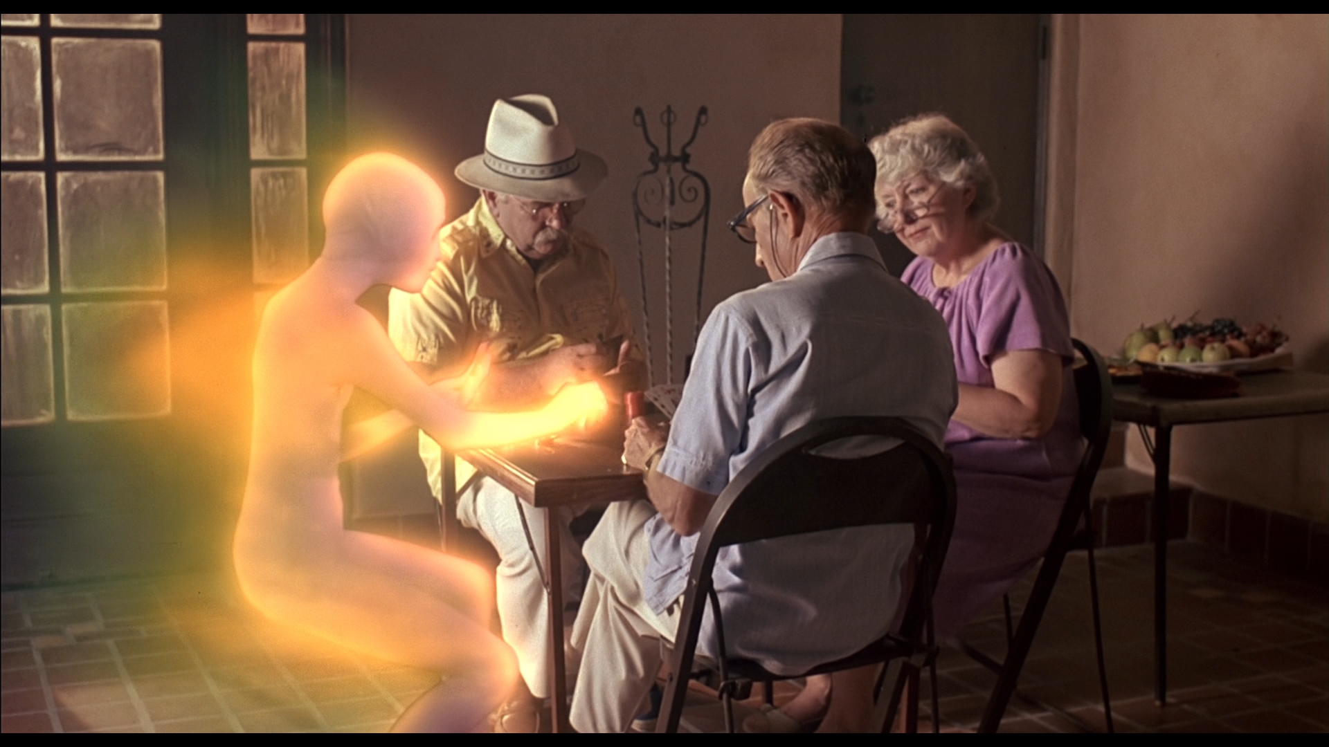 Screenshot from the movie Cocoon showing three elderly people sat at a table with a glowing golden orange alien.