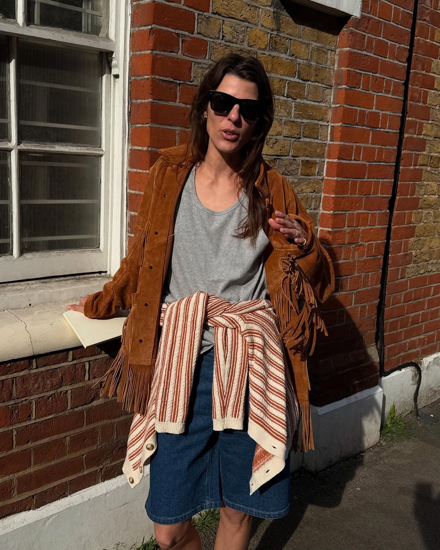 @lauravidrequin wearing a brown fringe jacket, gray top, and long shorts.