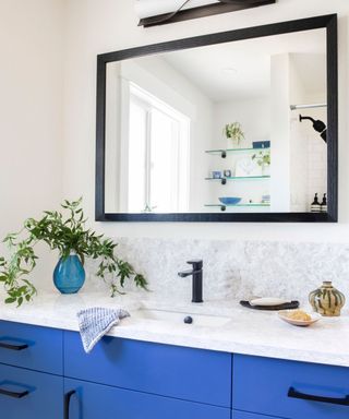 A bathroom sink area with a black framed mirror, a gray marble splashback, a sink with a black tap, a blue glass vase with long green foliage and a bright blue base with black handles
