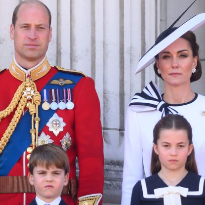 Prince William wears red military uniform and Kate Middleton wears a white dress with navy detailing, as they stand on the Buckingham Palace with their three kids, Prince George, Prince Louis, and Princess Charlotte
