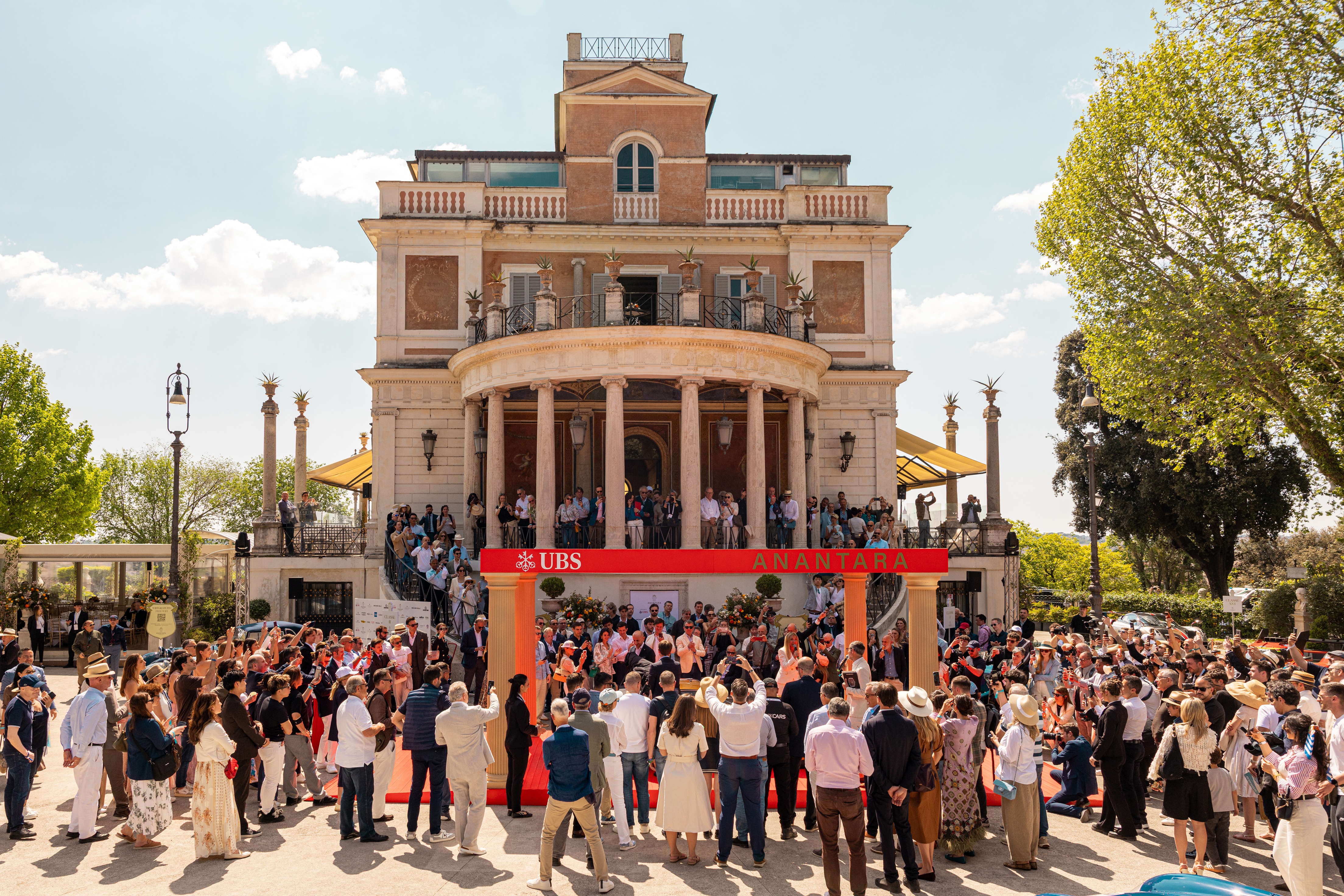 Crowds gather outside Casina Valadier in Rome for an Anantara Concorso Roma car unveiling beneath a red archway, with spectators filling the steps and balconies under a bright blue sky.