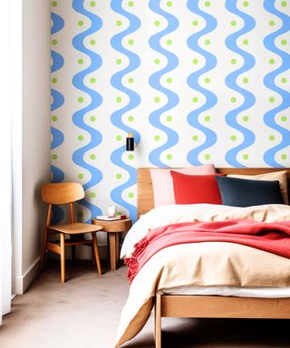 A wall with white and wavy cornflower blue stripes, a bed with light pink bedding and red throw pillows and blankets, with a wooden chair to the left of it