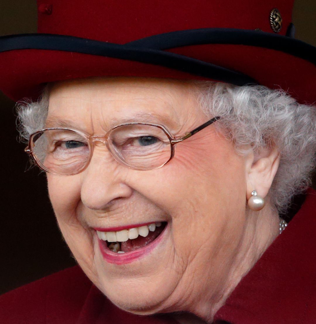 A headshot of Queen Elizabeth wearing a burgundy hat and coat while laughing 
