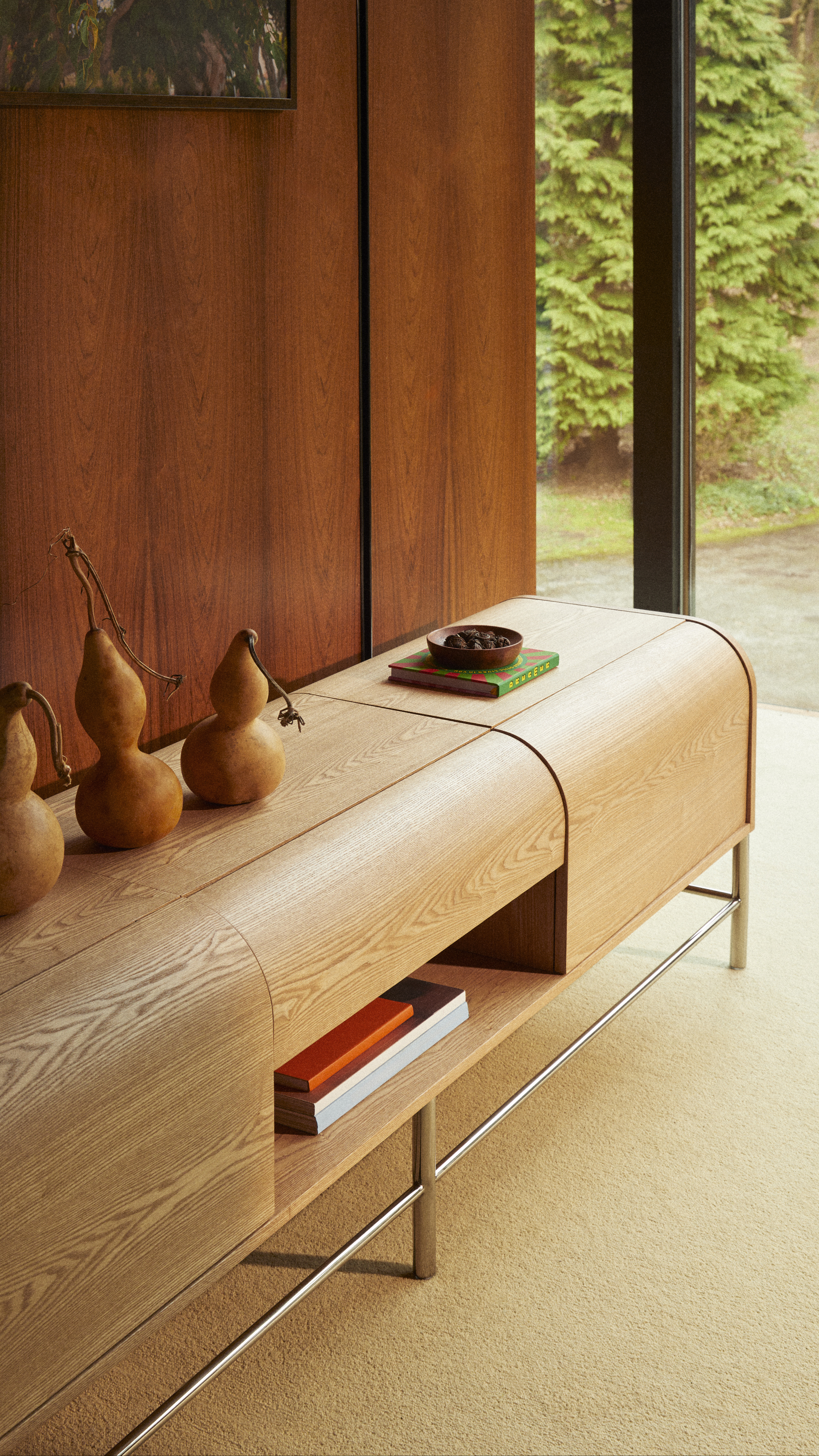 a curved timber sideboard with decorative pears on top, a timber panelled wall, beige carpets, and views of trees outside