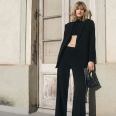 Young blonde woman wearing black tailored pants, blazer and crop top, with black handbag and rustic wall building her.