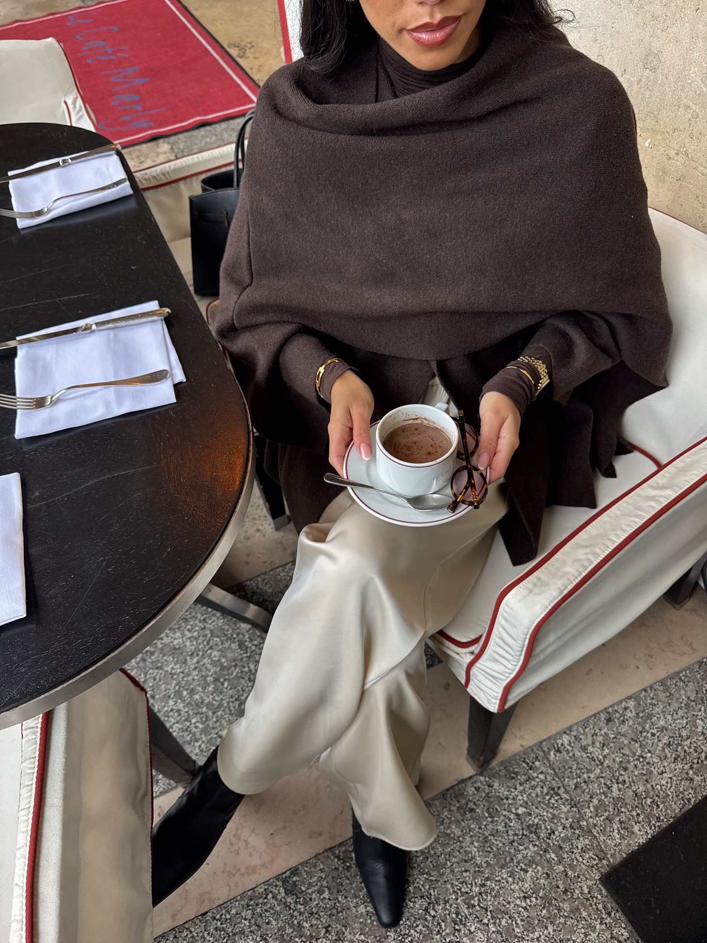Fashion person Olivia Orza shows what to wear in February. She wears cream satin trousers and a brown scarf coat. She finishes the look with black pointed toe boots and gold accessories over her cuffs. This image was recently taken from her Instagram.