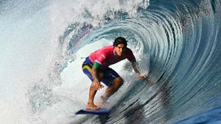 Brazil's Gabriel Medina rides in the barrel of a wave, in a red shirt and blue shorts on a blue surfboard, at the Paris 2024 Olympic Games, in Teahupo'o, Tahiti.