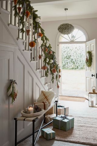 Scandi style Christmas entryway with foliage garland with ochre and white paper decorations, wreath on door, bench with tray, candle, paper decoration and gifts on the floor