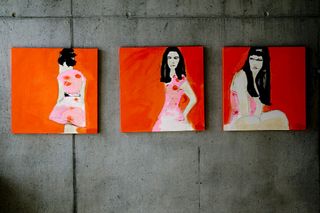 Three pieces of artwork with orange/red backgrounds, hang on a concrete wall