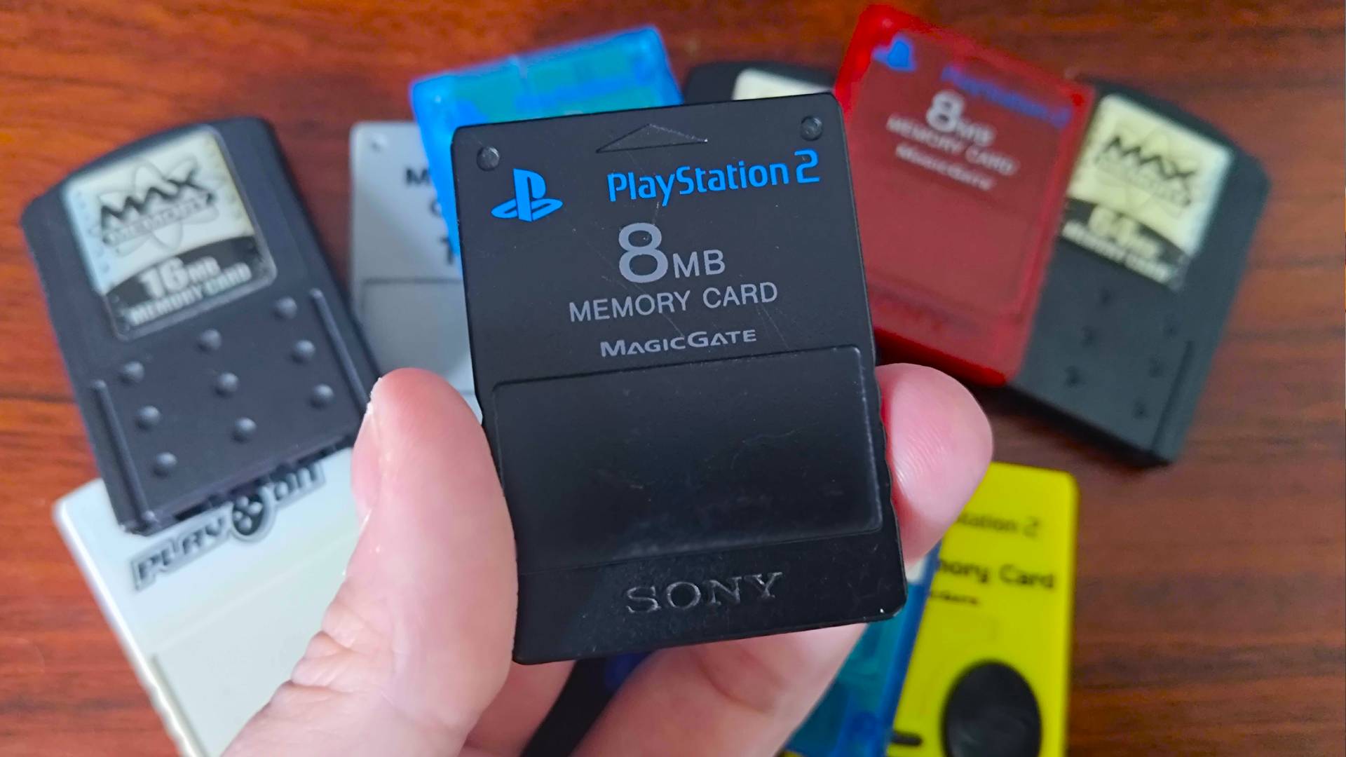 Hand holding PS2 memory card with various others in backdrop on desk.