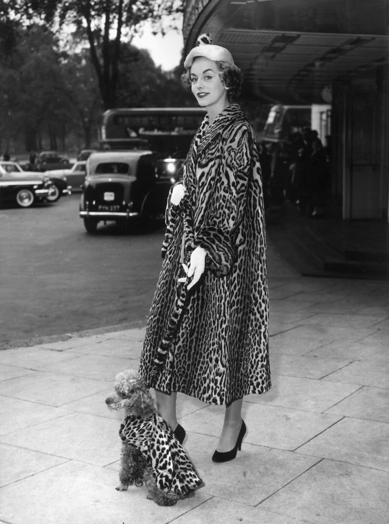 A fashionable woman wearing a leopard-print coat stands beside a toy poodle dressed in a matching leopard-print coat on a city street.