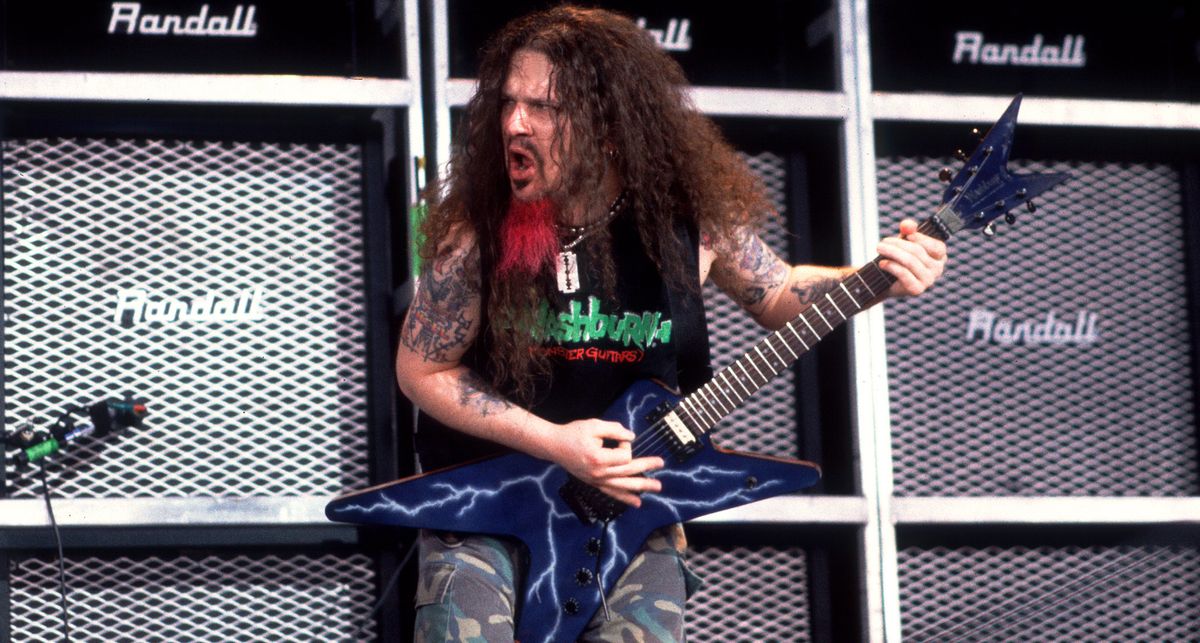 Dimebag Darrell riffs on his blue lightning graphic Washburn electric guitar in front of a wall of Randall guitar amps. He wears a Washburn T-shirt and his goatee is dyed red.