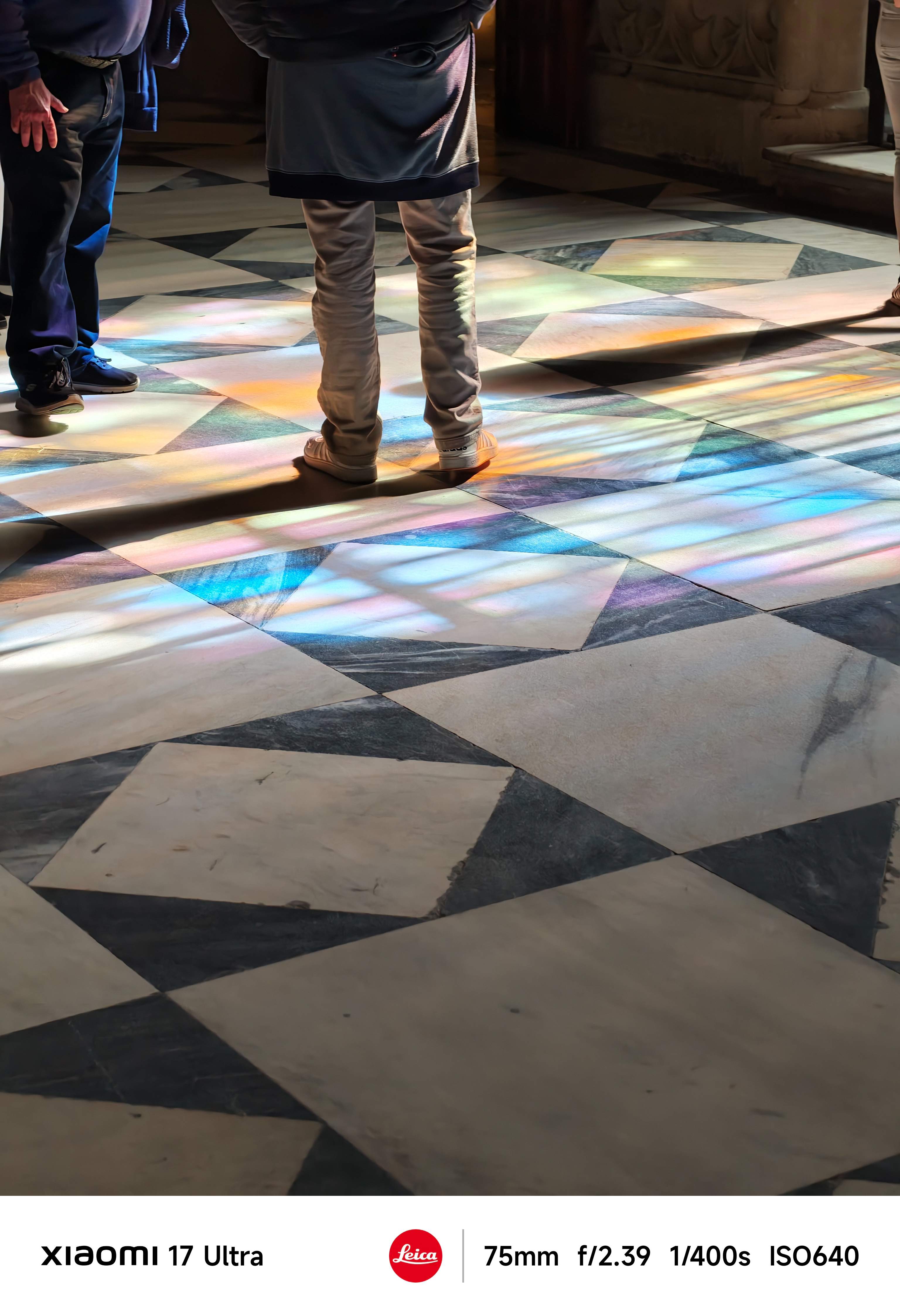 Lower view of people standing on a geometric tiled floor illuminated by colorful stained-glass light patterns indoors