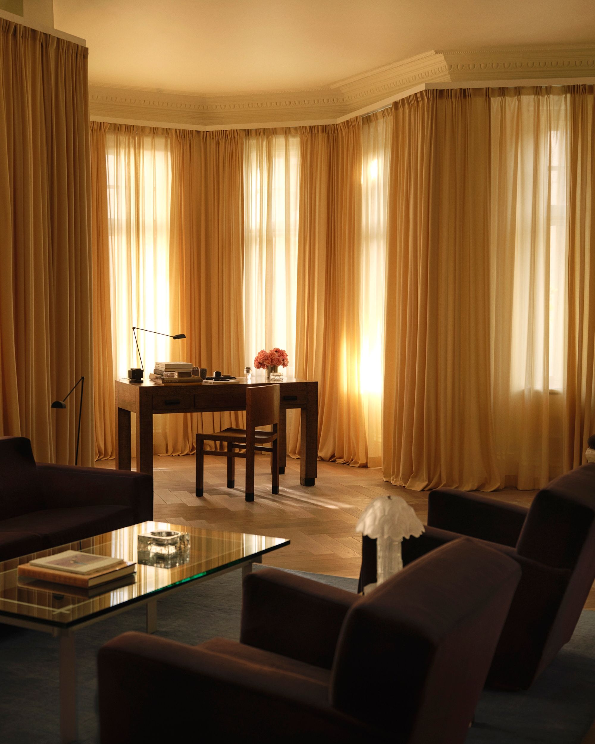 Image of a living room with sheer yellow curtains all around the windows of the room, with a desk in the bay window. There are two purple accent chairs and a matching sofa with a glass coffee table in between.