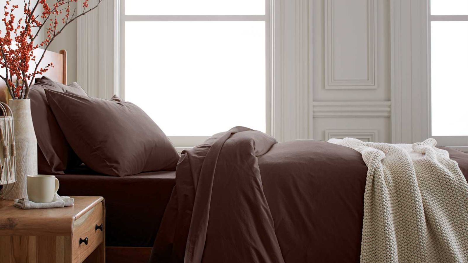 Dark brown bedding on a wooden bed with a wooden bedside table decorated with a vase of orange berries