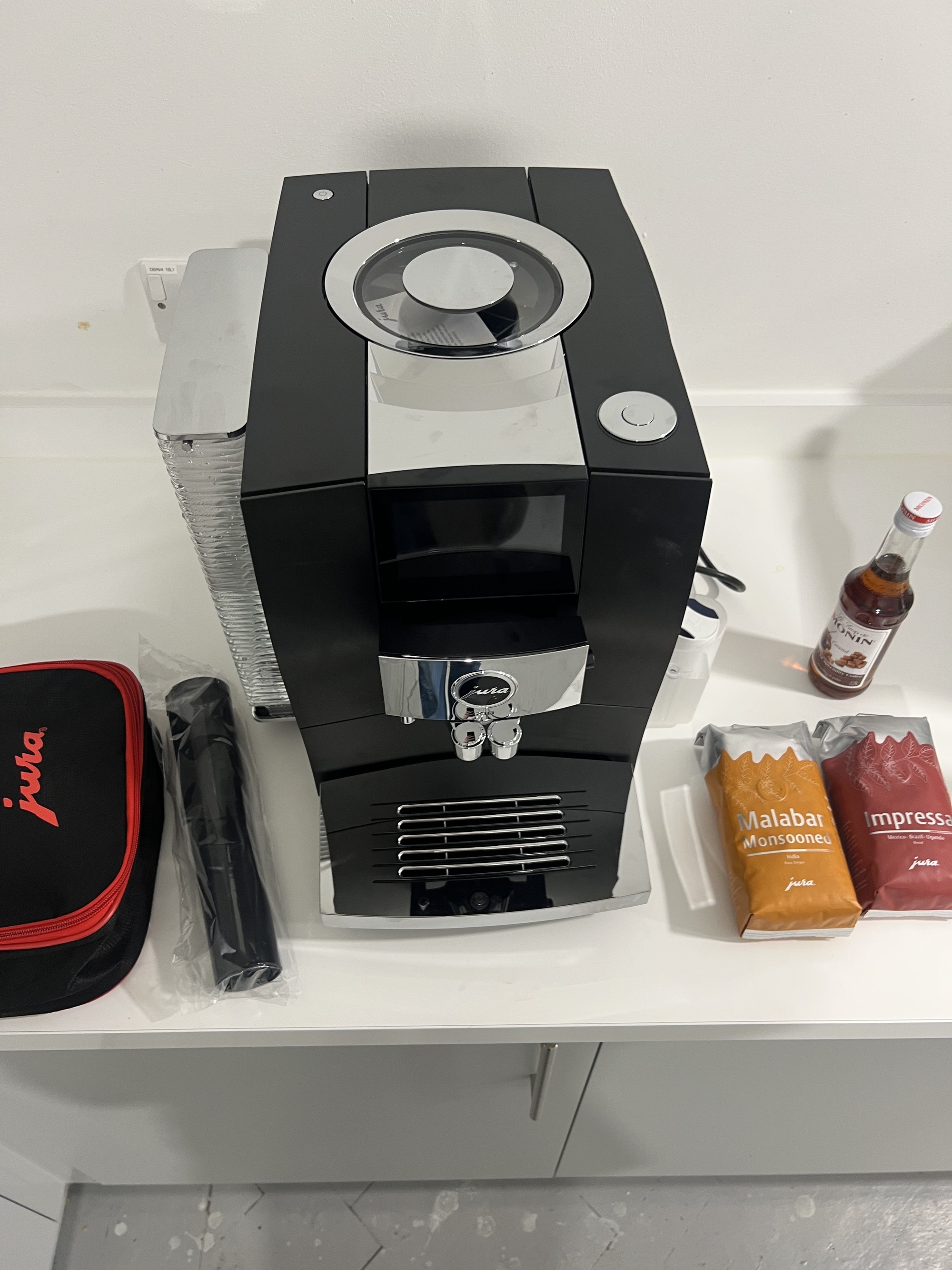 Image from above of a black and silver Jura Z10 coffee machine on a white kitchen countertop. There are two bags of coffee beans, a caramel syrup bottle, and Jura accessories also on the counter.