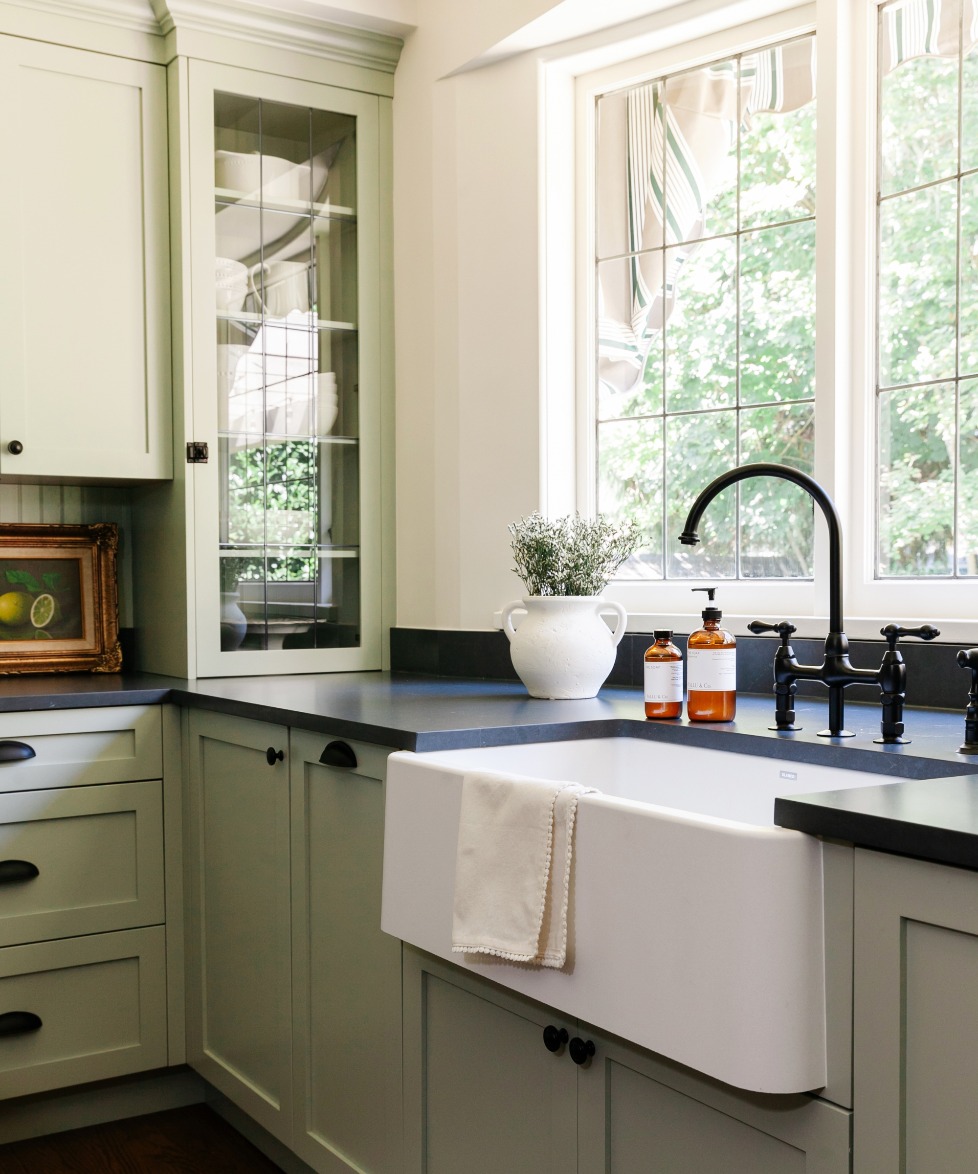 A sage green kitchen with large windows, a white basin sink, and a small plant sitting on a black countertop