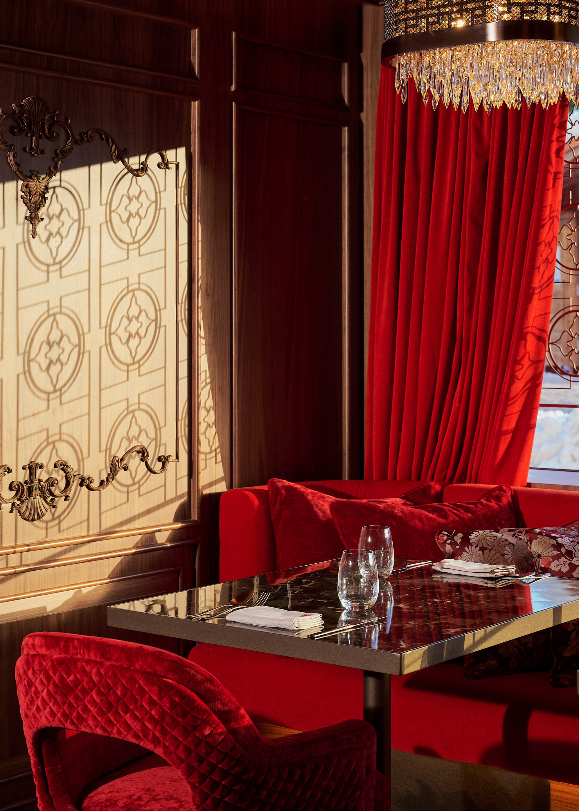A corner table in the bar at Maya Hotel Courchevel with a red sofa and dining chairs, red curtains behind, an opulent chandelier above the table, and the detailed window frame reflected on the back wall panel in the sunshine