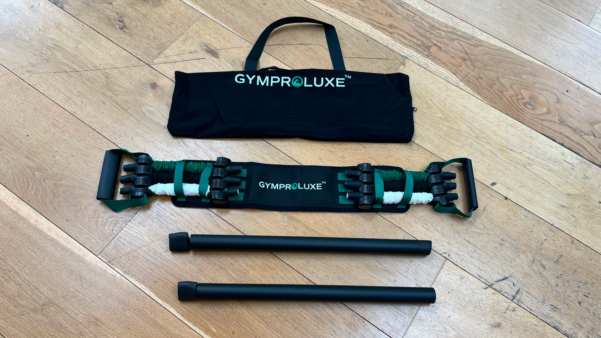 Fitness equipment arranged on wooden floor. At the top is a black carry bag with the work Gymproluxe printed in white text. Underneath is a belt with white, black and green tubes attaching the belt with two handles either end. Underneath that is two black bars.