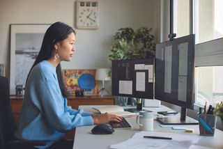 A focused woman in a blue sweater works at a desk with dual monitors. She is typing, surrounded by office decor, a clock, and natural light from a window.