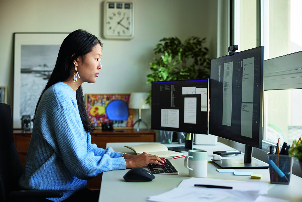 A focused woman in a blue sweater works at a desk with dual monitors. She is typing, surrounded by office decor, a clock, and natural light from a window.
