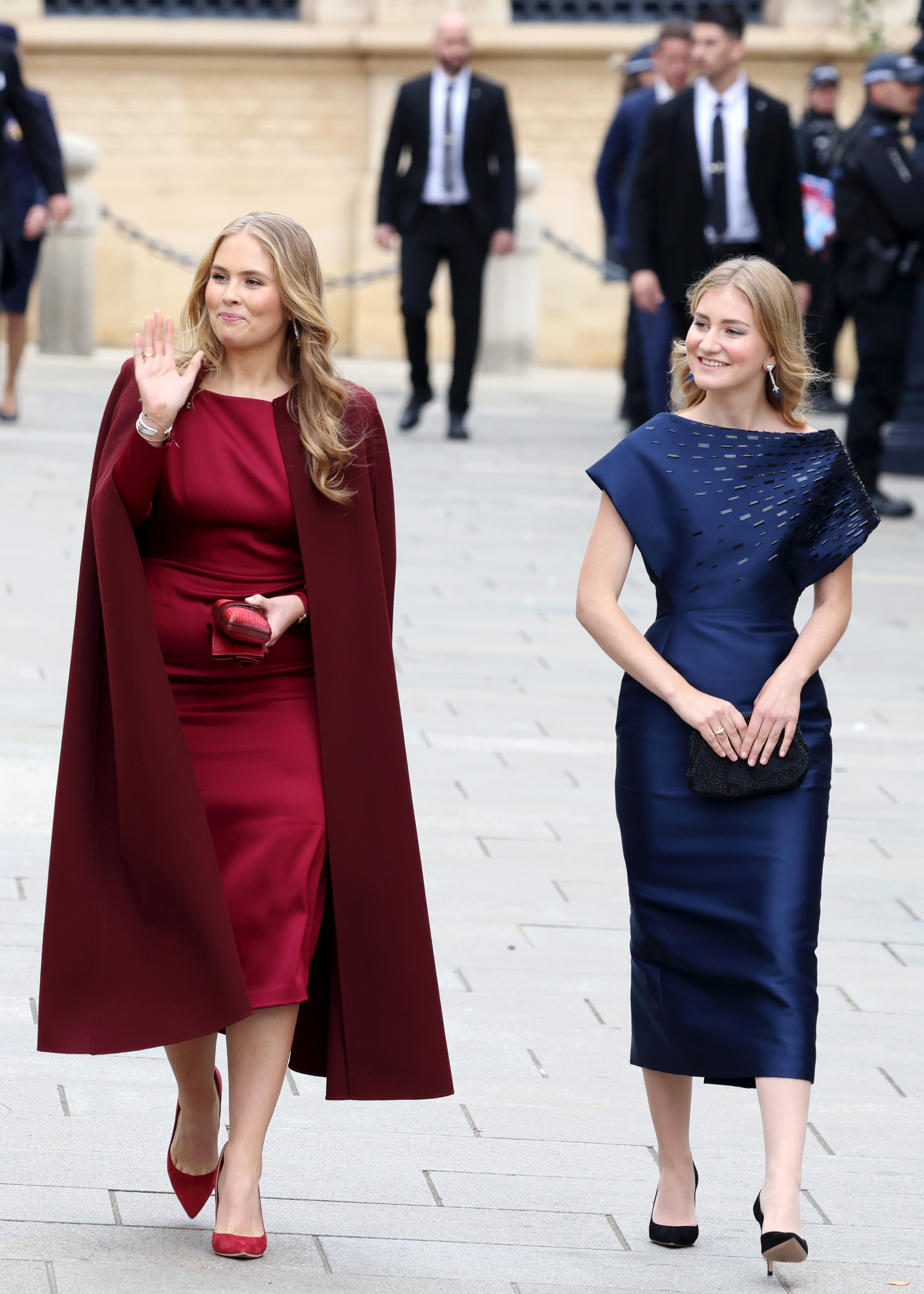 Princess Catharina-Amalia wearing a maroon dress and waving walking next to Princess Elisabeth in a navy dress