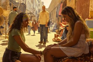 Kiara (Madison Bailey) kneels in front of Sarah (Madelyn Cline) with her hand on her knee, with Rafe (Drew Starkey) standing in the background, in episode 410 of Outer Banks.