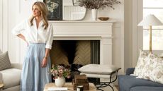 mcgee & co founder designer shea mcgee photographed stood beside a white fireplace in a neutral living room with a navy couch wearing a white shirt and blue and white stripe maxi skirt