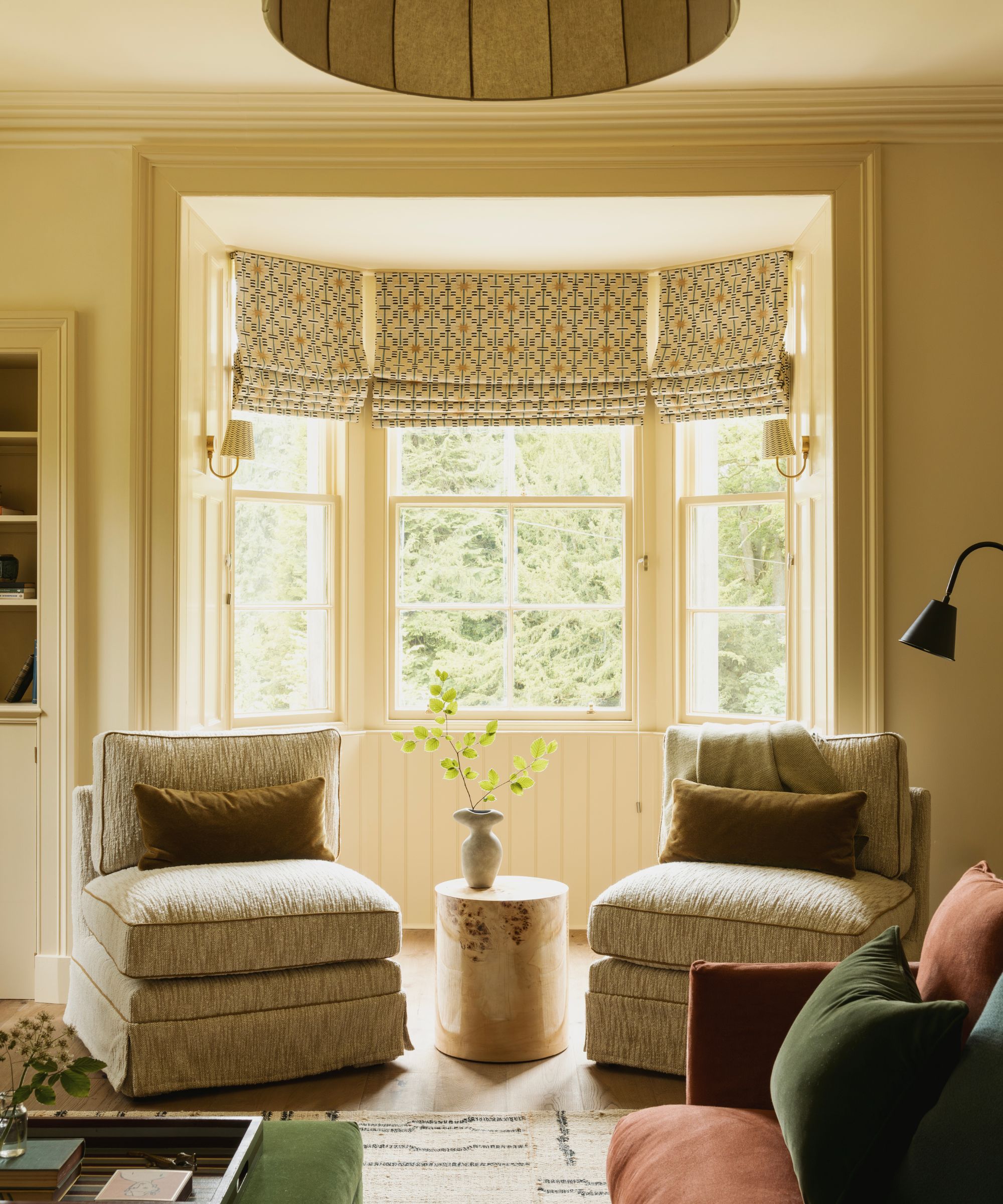 Butter yellow living room with large bay window and patterned Roman blinds, two slipper chairs with velvet cushions on top, a rust velvet sofa, a Berber rug, and a coffee table in the centre.